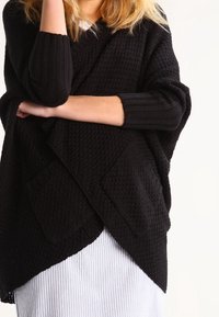 Žena v černém strukturovaném pleteném kardiganu s kapsami, přes světle pruhovanou sukni, se zavřenýma rukama, na bílém pozadí.