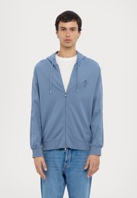 Világoskék, cipzáros kapucnis pulóver puha anyagból, húzózsinórral, két elülső zsebbel és az "Armani Exchange" márkázással az ujjakon.