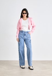Lys rosa cropped hettegenser, hvit tettsittende topp, høyhuskede lyseblå cargo-jeans med store lommer, og svarte solbriller. Klumpete sko fullfører looken.