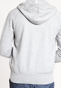 Persona que lleva una sudadera con capucha de color gris claro vista desde atrás, con las mangas ligeramente arrugadas, combinada con pantalones oscuros.