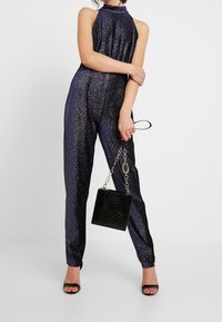 Mono azul marino con cuello halter, que presenta una textura metálica brillante. Combinado con un bolso negro con estampado de cocodrilo y tacones abiertos negros.