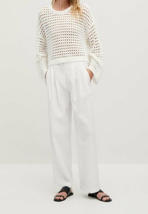 White crochet sweater with wide sleeves paired with high-waisted white trousers and black slide sandals, set against a neutral background.