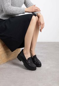 Mocasines de cuero negro con parte superior texturizada, tachuelas de metal, suelas de plataforma gruesas y un acabado suave, combinados con una falda negra y un suéter gris.