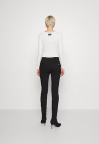 Witte geribbelde lange mouwen top gecombineerd met zwarte strakke broek en zwarte stiletto hakken. Opvallend label zichtbaar aan de achterkant.