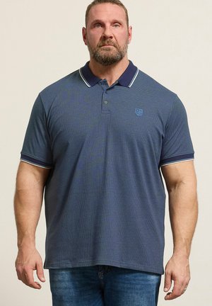 Brodaty mężczyzna w granatowej koszulce polo z białym paskowanym kołnierzykiem i rękawami, stojący na jasnym, jednolitym tle.