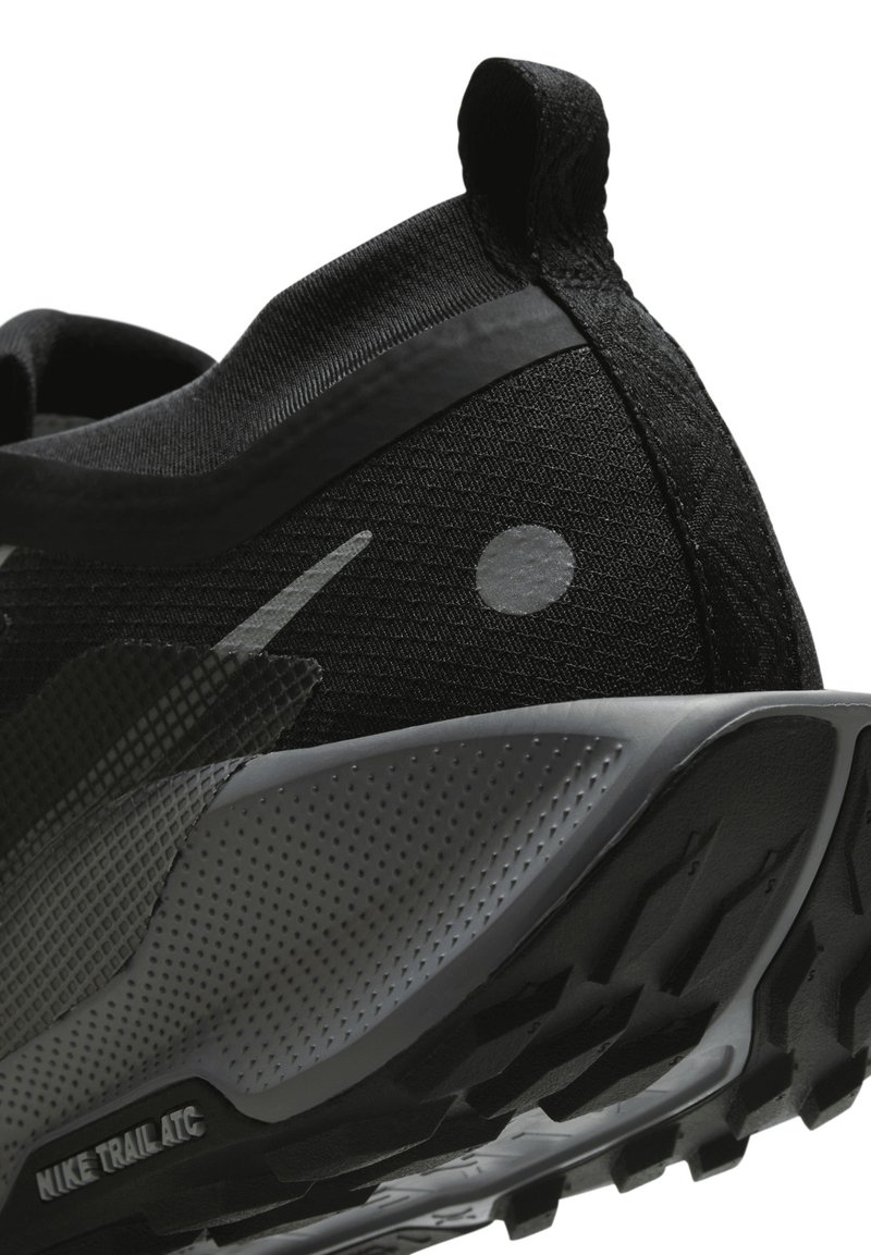 Zapatilla deportiva negra con parte superior de malla texturada, acentos grises, un talón redondeado y suela de goma con patrón para mayor tracción. Marca Nike Trail ATC visible.