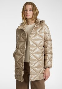 Beige pufferjack met een geometrisch gewatteerd patroon en een capuchon. Heeft drukknoopjes en zijzakken. Draag over een licht beige trui.