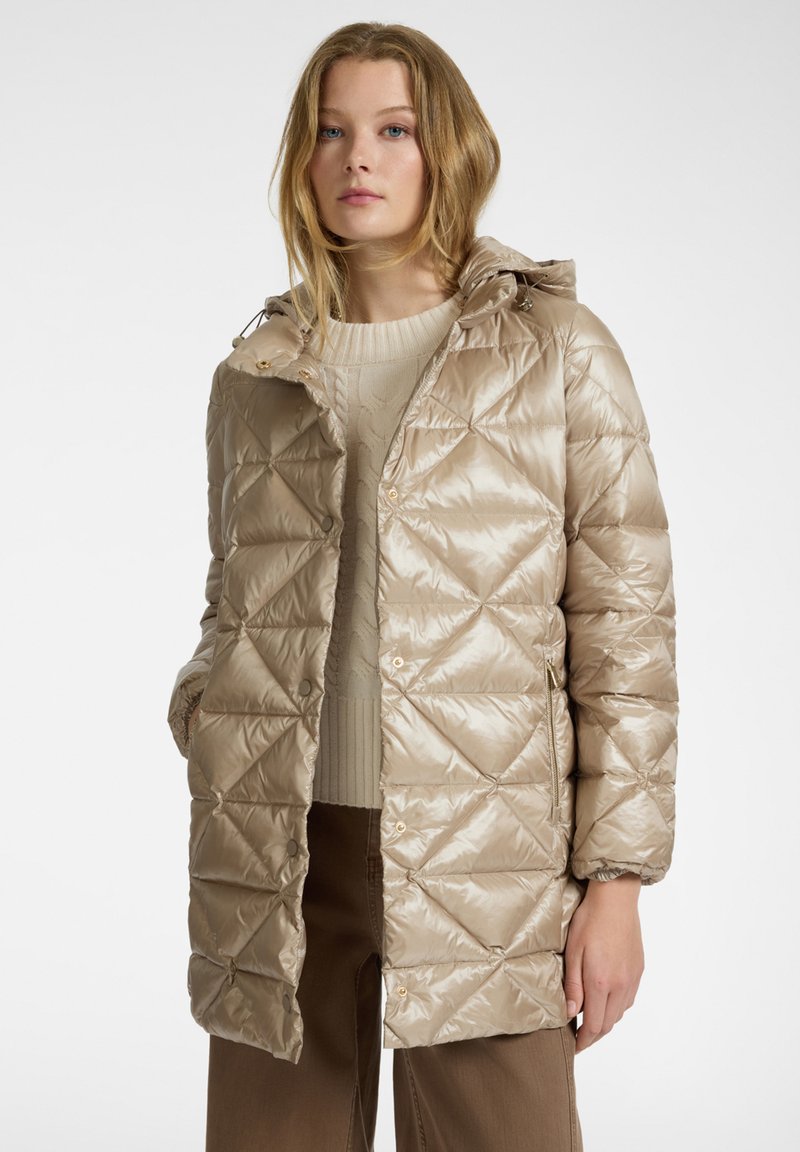 Beige pufferjack met een geometrisch gewatteerd patroon en een capuchon. Heeft drukknoopjes en zijzakken. Draag over een licht beige trui.