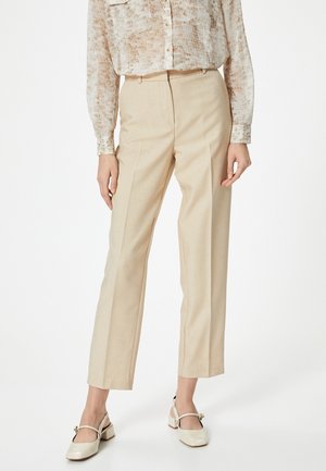 Beige tailored trousers with straight legs, made from lightweight fabric. The model wears a patterned sheer top and light-colored chunky heels.