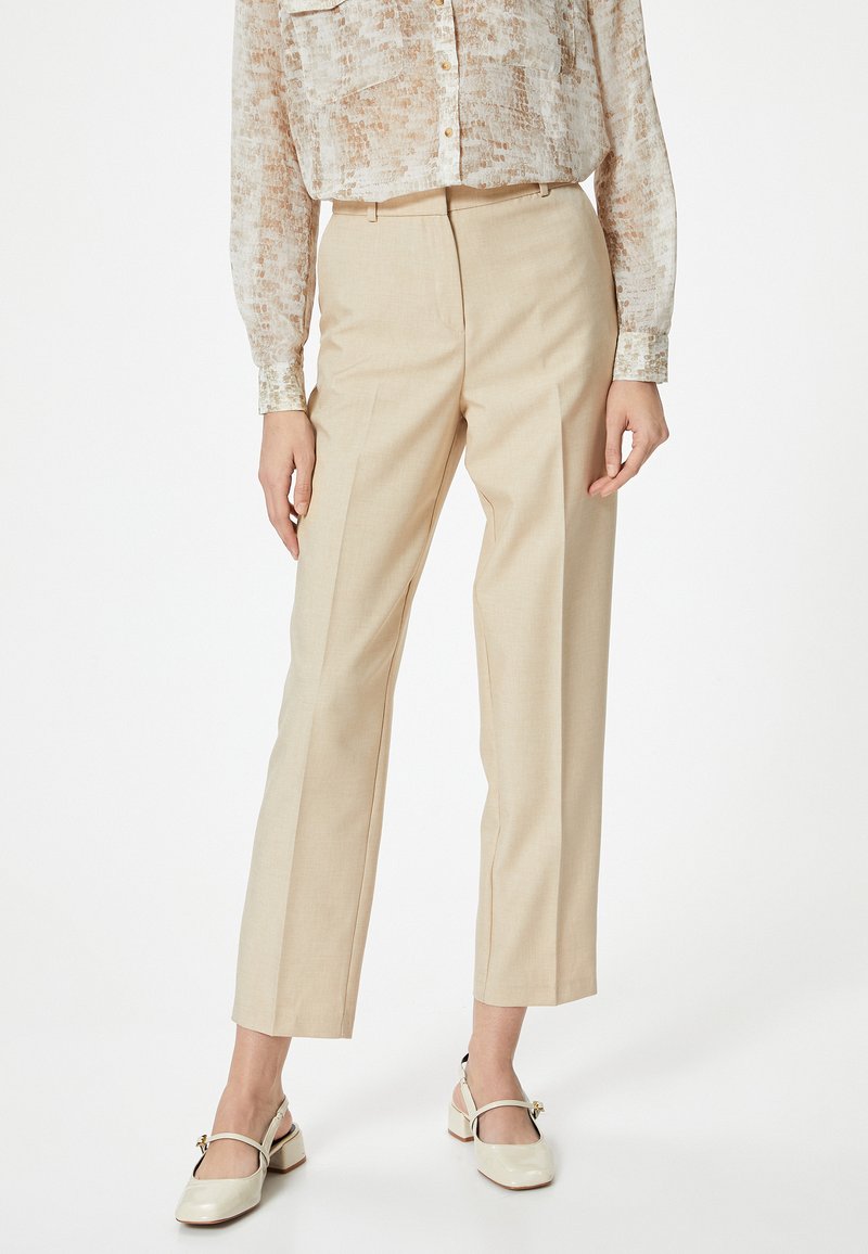 Beige op maat gemaakte broek met rechte pijpen, gemaakt van lichtgewicht stof. Het model draagt een patroon met een doorschijnende top en lichtgekleurde stevige hakken.