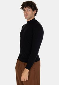 Maglione a collo alto nero in un design aderente, texture in maglia, maniche lunghe, con una finitura liscia e senza motivi visibili. Abbinate a pantaloni marroni.