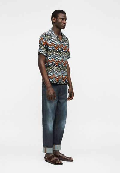 Άνδρας στέκεται φορώντας πολύχρωμο πουκάμισο με μοτίβο ζιγκ-ζαγκ και κοντά μανίκια, ξεθωριασμένα μπλε τζιν με γυρισμένα μανίκια και καφέ δερμάτινα σανδάλια.