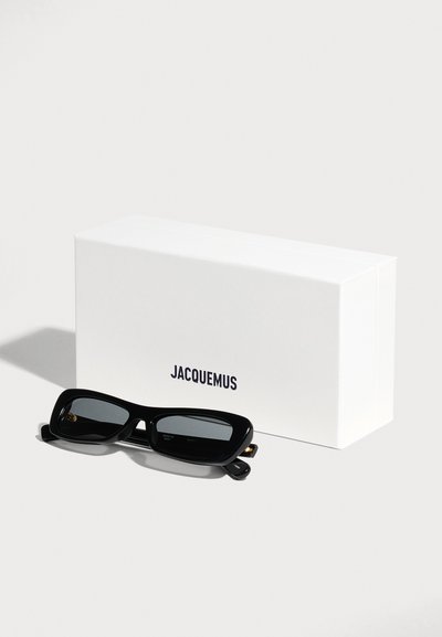 Lunettes de soleil rectangulaires noires reposant devant une boîte blanche de la marque Jacquemus sur une surface claire.