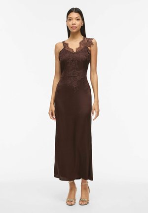 Mujer que lleva un vestido sin mangas de satén marrón oscuro con detalles de encaje en el cuerpo y los hombros, combinado con tacones abiertos dorados.