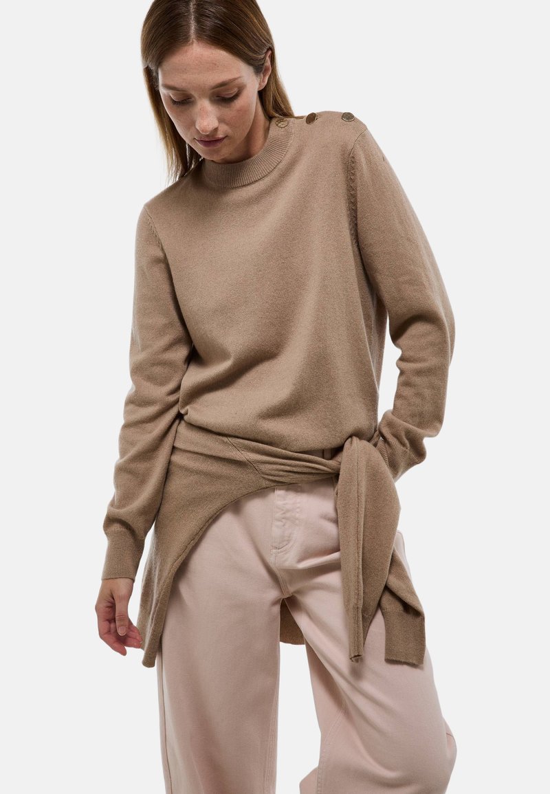 Femme portant un pull beige à manches longues avec des boutons sur l'épaule et un pantalon rose clair, posant avec une main dans la poche.