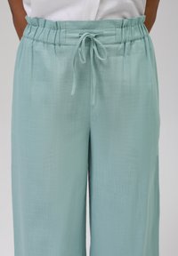 Pantalones de pierna ancha en azul claro, fabricados con un tejido texturizado. Presentan una cinturilla fruncida con cordón ajustable. Diseño casual.