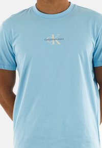 Camiseta de algodón azul claro con cuello redondo, mangas cortas y un logo impreso central en crema y gris: "Calvin Klein Jeans."