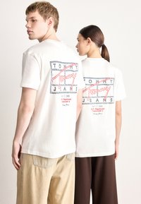 Valkoisissa puuvillaisissa T-paidoissa on takana punaisia ja sinisiä graafisia kuvioita, joissa lukee "TOMMY" ja "JANS." Mallit käyttävät vaaleita ja tummia housuja.