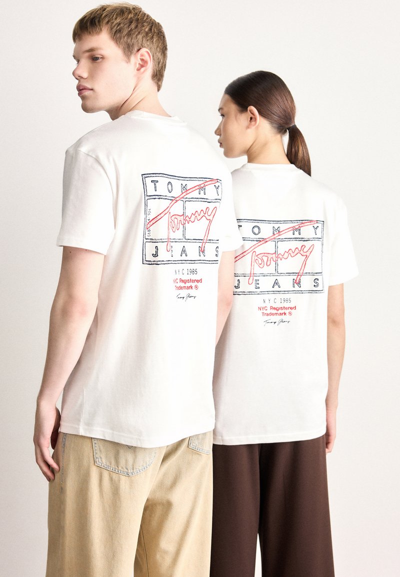 Valkoisissa puuvillaisissa T-paidoissa on takana punaisia ja sinisiä graafisia kuvioita, joissa lukee "TOMMY" ja "JANS." Mallit käyttävät vaaleita ja tummia housuja.