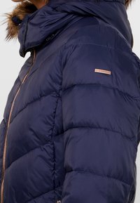 Tmavě modrá péřová bunda se zipovým zapínáním, kapucí a umělým kožešinovým lemováním. Má prošité panely a kovovou značku na rukávu.