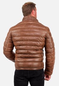 Giacca di pelle marrone con design trapuntato e colletto rialzato; presenta una texture liscia e una silhouette aderente, indossata con jeans neri.