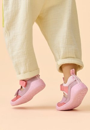 Child wearing cream pants and pink soft sole shoes, standing on tiptoes against beige background.