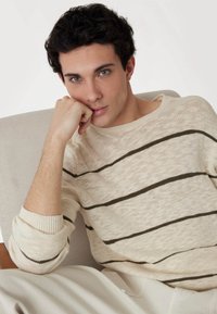 Pull en maille beige avec des rayures vert foncé, poignets côtelés. Le modèle repose son menton sur sa main, assis sur une chaise de couleur claire devant un fond neutre.