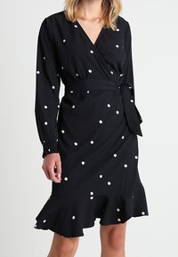 Robe portefeuille noire à pois blancs, manches longues avec poignets froncés, taille ceinturée et ourlet à volants. Fabriquée en tissu léger.