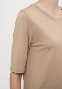Beige krátký rukáv top vyrobený z měkké, strukturované tkaniny. Má kulatý výstřih a volný střih, s viditelnými švy na rukávech.