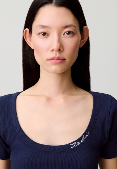 Top corto azul marino con mangas cortas acanaladas, cuello redondo y bordado con la palabra "Claudie" en blanco en el lado izquierdo cerca del cuello.