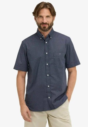 Homme portant une chemise à manches courtes bleu marine à boutons avec des points blancs et un pantalon beige, debout avec une main dans la poche.