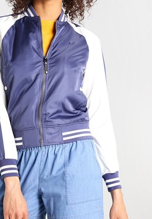 Personne portant une veste universitaire bleu et blanc par-dessus un haut jaune et un short bleu clair à taille élastique sur un fond uni.