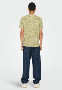 Camiseta de manga corta color oliva con un patrón floral, combinada con unos vaqueros oscuros de corte holgado y zapatos de color claro.