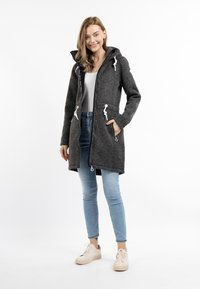 Grauer, kapuzenförmiger, langer Mantel mit Reißverschluss, weißen Kordeln und Taschen, kombiniert mit hellblauen Jeans und beigen Sneakers.