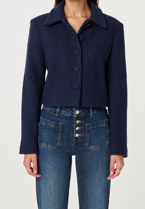 Mujer que lleva una chaqueta corta azul marino con botones y vaqueros azules de tiro alto con cuatro botones plateados y bolsillos delanteros con parche.