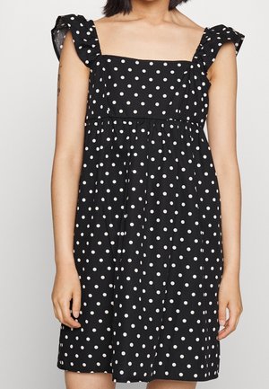Kobieta ubrana w czarną sukienkę bez rękawów w białe grochy z falbanianymi ramiączkami, stojąca na jednolitym tle.