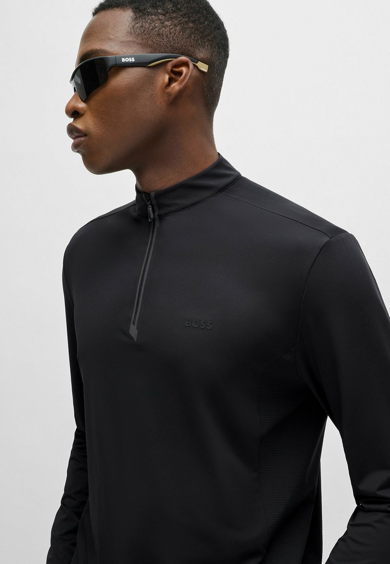 Camiseta de rendimiento negra de manga larga con cuello de media cremallera, que presenta un acento texturizado y un logo de la marca en tono sobre el pecho.