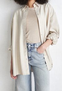 Camicia button-up beige a righe, vestibilità rilassata, tessuto leggero. Abbinata a una t-shirt beige e jeans in denim a vita alta di un lavaggio chiaro.