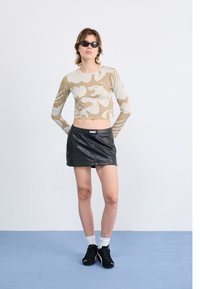 Beige long-sleeve crop top with abstract print, paired with a black leather mini skirt. Model wears black sunglasses and black sneakers.