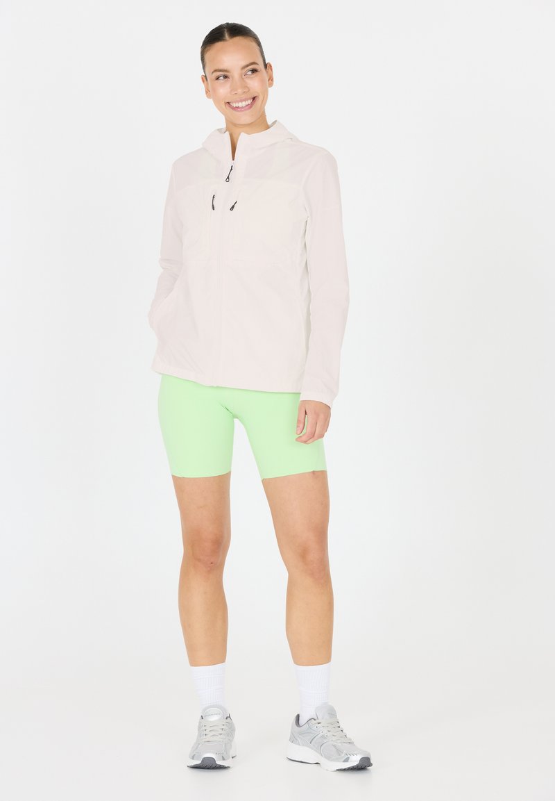 Lätt, ljusrosa zip-jacka med huva, kombinerad med klara gröna shorts. Modellen har på sig grå träningsskor och vita strumpor.