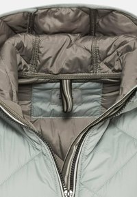 Veste matelassée gris clair avec un extérieur lisse, dotée d'une fermeture éclair, d'une doublure intérieure en brun sobre et d'une étiquette de logo à l'intérieur.