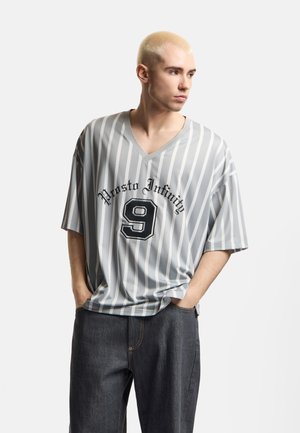 Młoda osoba z wybielonymi krótkimi włosami, ubrana w luźną szaro-białą koszulkę w paski z dekoltem w serek i napisem „Prosto Infinity 9”, z rękami w kieszeniach.