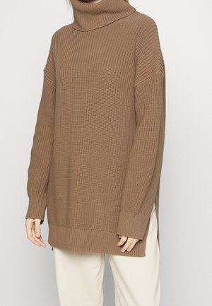 Persoon die een losse, bruine gebreide coltrui met lange mouwen en crèmekleurige broek draagt tegen een effen achtergrond.