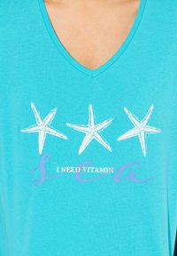 T-shirt in cotone turchese con scollo a V che presenta tre stelle marine bianche e la frase "HO BISOGNO DI VITAMINA MARE" in caratteri viola.