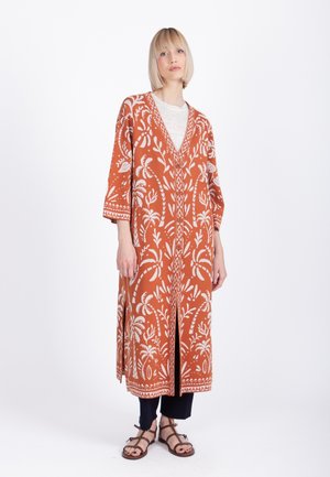 Mujer con bob rubio que lleva un cárdigan largo color óxido con patrones florales blancos, pantalones negros y sandalias marrones, de pie contra un fondo blanco.