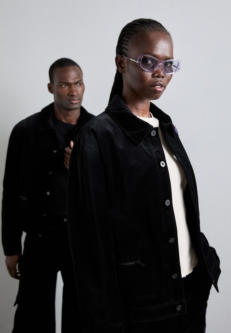 Schwarze Cord-Jacke mit Knopfverschluss und seitlichen Taschen, getragen über einem hellen Hemd; zeigt ein Model mit lila eckigen Sonnenbrillen.