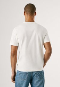 Camiseta blanca de manga corta con cuello redondo, tejido suave y una pequeña etiqueta con el logo en el costado. Se combina con jeans de denim azul.