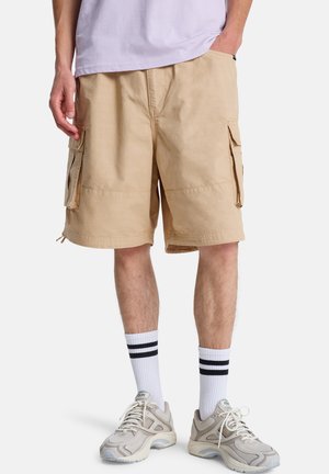 Beige Cargo-Shorts mit zwei Seitentaschen, einem lockeren Schnitt und Steppnähten. Kombiniert mit weißen Socken mit schwarzen Streifen und sportlichen Sneakers.