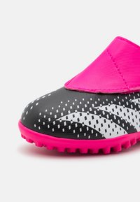 Zapato de fútbol negro con superficie texturizada, cinta elástica rosa, acentos de rayas blancas y una suela de goma rosa brillante con taquetes de tracción.