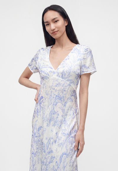 Jeune femme aux longs cheveux foncés portant une robe blanche à motifs floraux bleus, debout avec une main sur la hanche devant un fond uni.